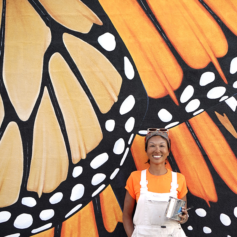 Jane Kim, Ink Dwell studio in front of one of a butterfly mural